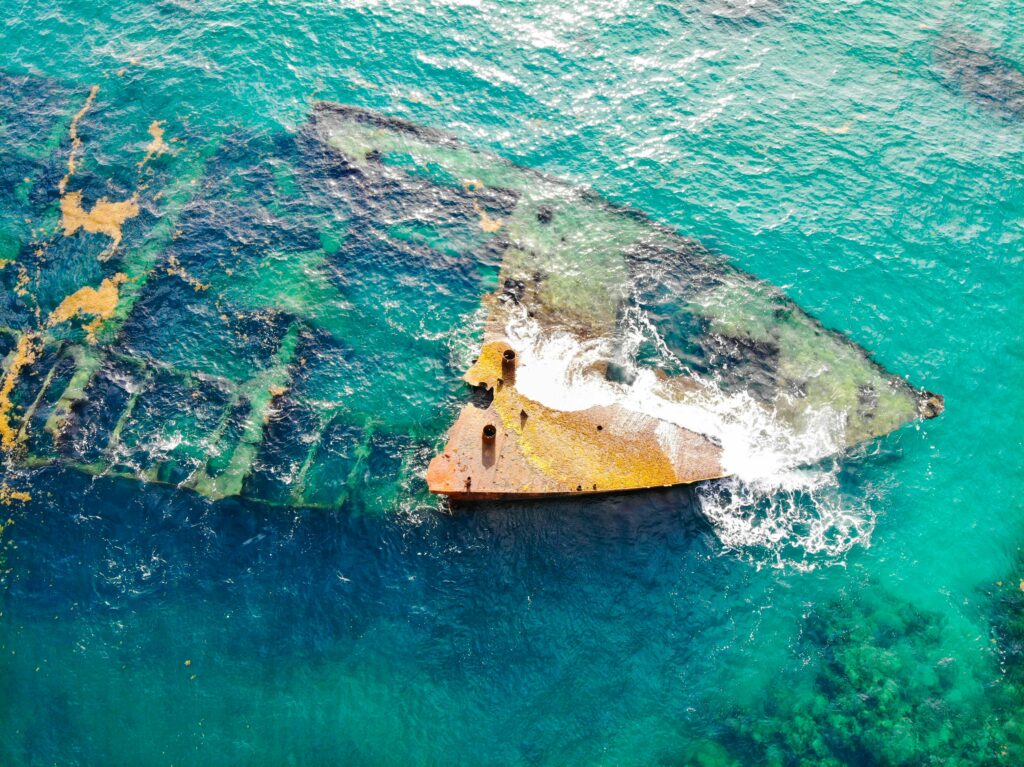 Drone shot capturing a sunken shipwreck in vibrant turquoise waters, surrounded by waves and marine life.