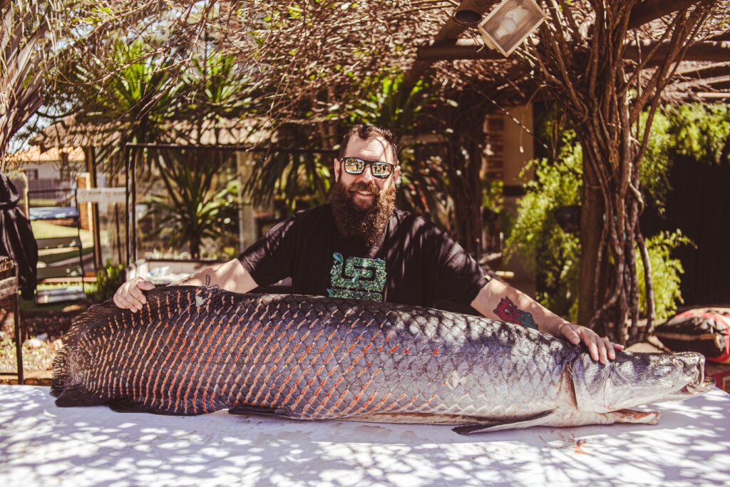 Man with beard outdoors displaying a giant fish on a table, captured in a sunlit setting.